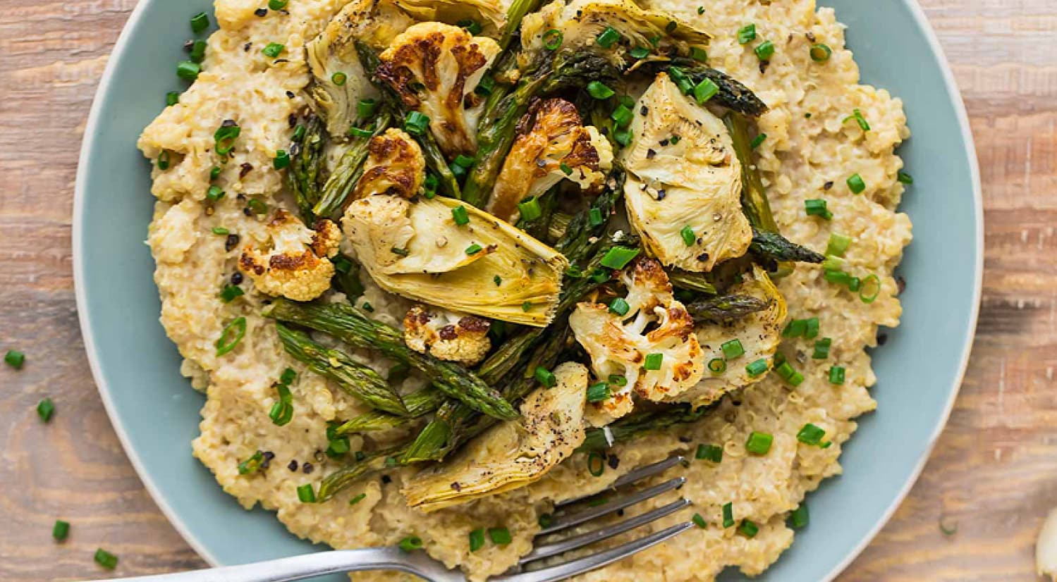 A plate of creamy risotto topped with roasted asparagus, cauliflower, and artichoke pieces, garnished with chopped chives. A fork rests on the plate.