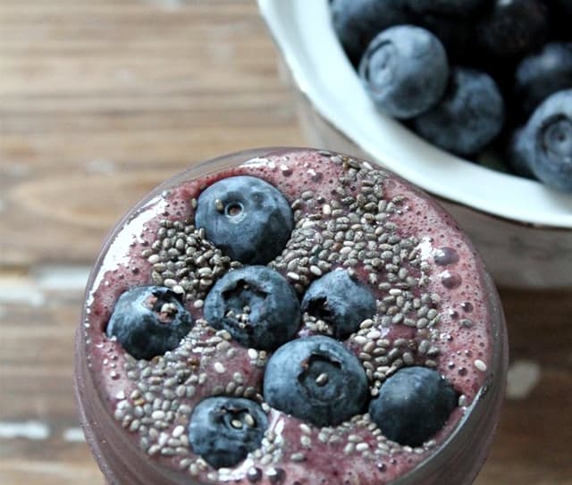 A close-up of a smoothie topped with fresh blueberries and chia seeds, with a bowl of blueberries in the background on a wooden surface.