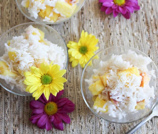 Three glass bowls of rice pudding with fruit and shredded coconut, placed on a woven surface, with yellow and purple flowers as decoration.