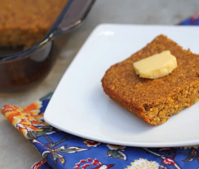 A slice of cornbread with a pat of butter on top sits on a white plate next to a glass baking dish, placed on a colorful floral napkin.