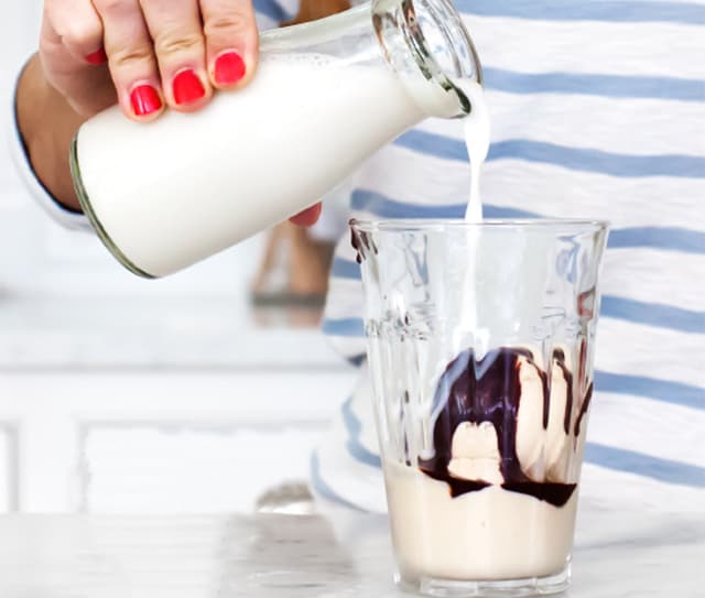 A person pours milk from a glass bottle into a glass containing ice cream and chocolate syrup.