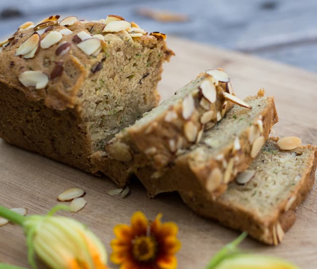 A loaf of sliced almond zucchini bread sits on a wooden cutting board, garnished with sliced almonds. Flowers are in the foreground.