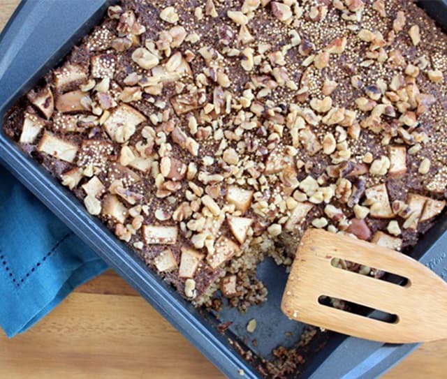A square baking pan with a baked dessert topped with chopped nuts and fruit pieces, with a wooden spatula and a missing corner piece.