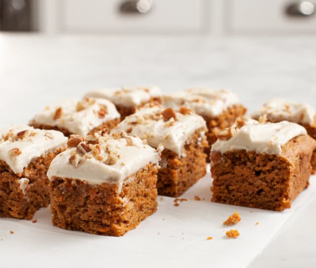 Eight frosted carrot cake squares topped with chopped nuts are arranged on a white surface with a blurred kitchen background.