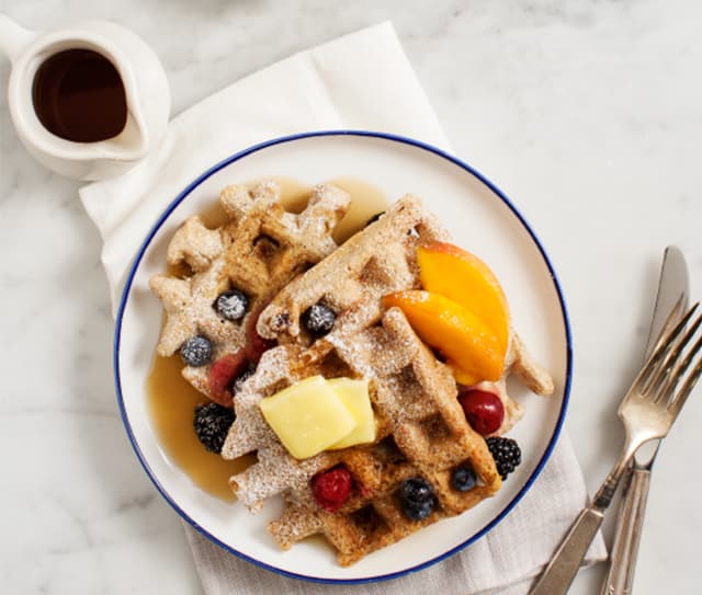 Plate of waffles topped with butter, powdered sugar, blueberries, blackberries, raspberries, peach slices, and syrup, with a fork, knife, and a small pitcher of syrup on the side.