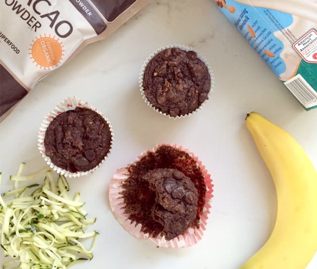 Three chocolate muffins on a white surface with a banana, shredded zucchini, a bag of cacao powder, and a carton of eggs nearby.