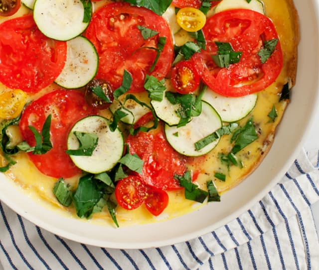 A baked frittata topped with sliced tomatoes, zucchini, cherry tomatoes, and chopped fresh herbs in a white dish on a striped cloth.