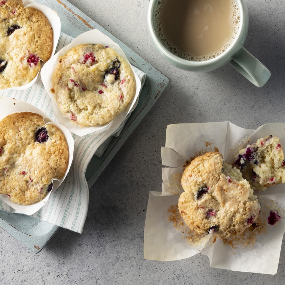 Four berry muffins on a tray with a striped napkin beside a cup of coffee on a gray surface; one muffin is broken open on parchment paper.