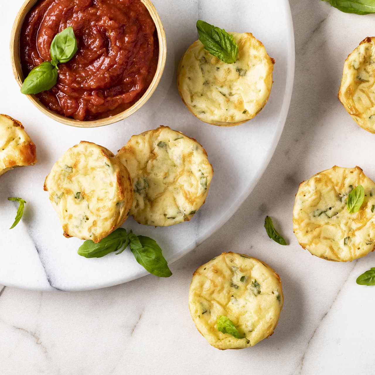 Six mini egg muffins with herbs are arranged on a white surface with a bowl of tomato sauce and fresh basil leaves for garnish.