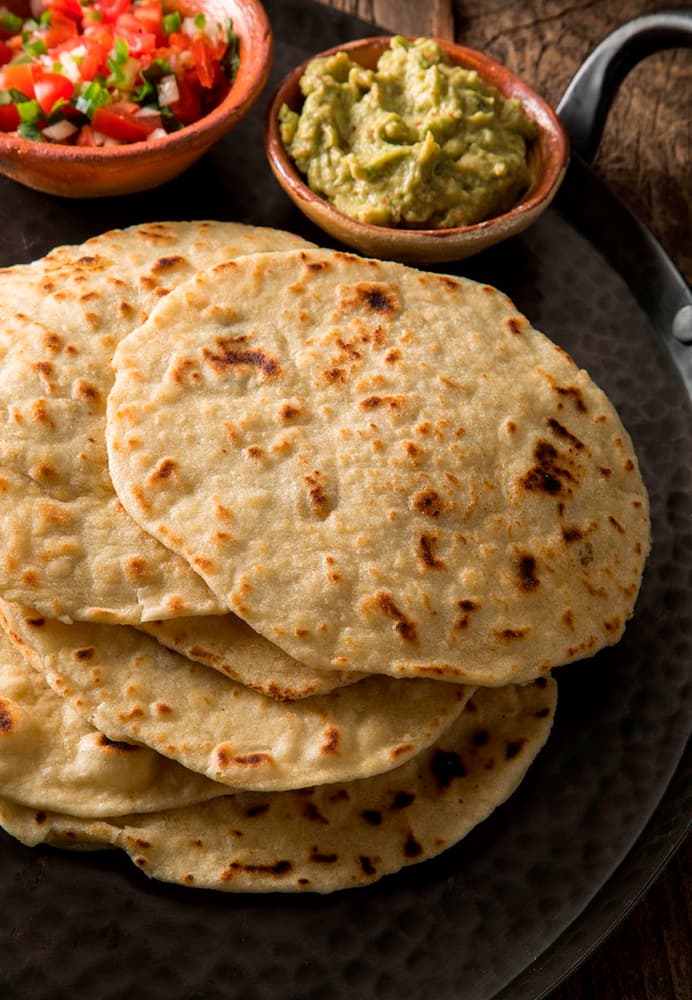 A plate with several stacked flour tortillas, with small bowls of guacamole and salsa on the side.
