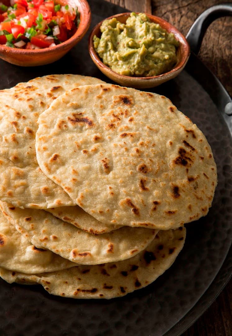 A plate with several stacked flour tortillas, with small bowls of guacamole and salsa on the side.