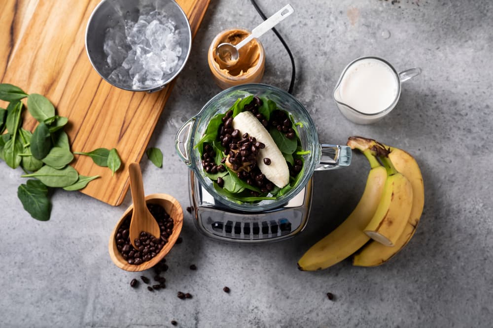 Overhead view of a blender with spinach, banana, and coffee beans, surrounded by bananas, spinach, ice, peanut butter, and milk on a kitchen counter.
