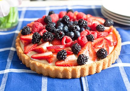 A fruit tart with a pastry crust, topped with strawberries, blackberries, raspberries, and blueberries, is displayed on a blue checkered tablecloth.
