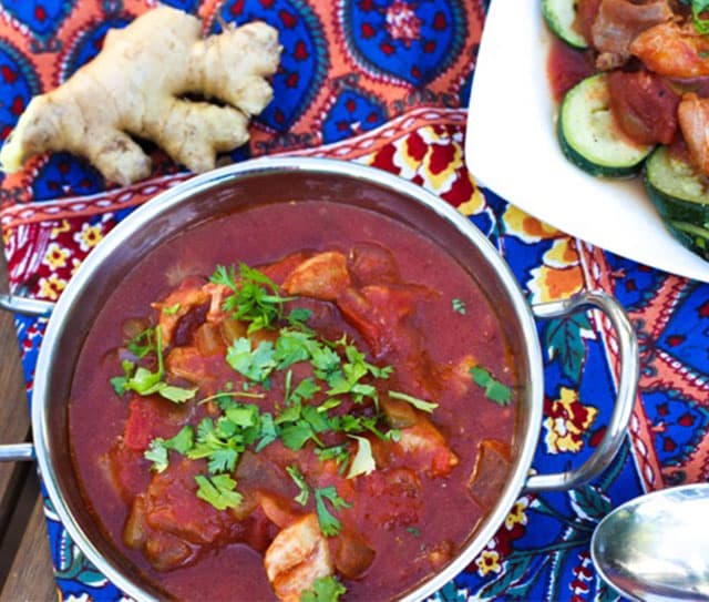 A bowl of chicken curry garnished with cilantro, beside sliced zucchini and a piece of ginger, on a colorful patterned tablecloth.