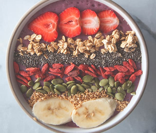 A smoothie bowl topped with sliced strawberries, granola, chia seeds, goji berries, pumpkin seeds, flaxseeds, and banana slices, arranged in neat rows.