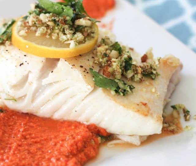A piece of baked white fish topped with a lemon slice and herb crust, served with a red sauce on a white plate.