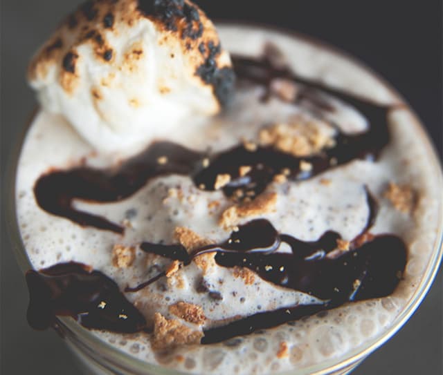 A close-up of a frothy drink topped with chocolate drizzle, crumbled graham crackers, and a toasted marshmallow.