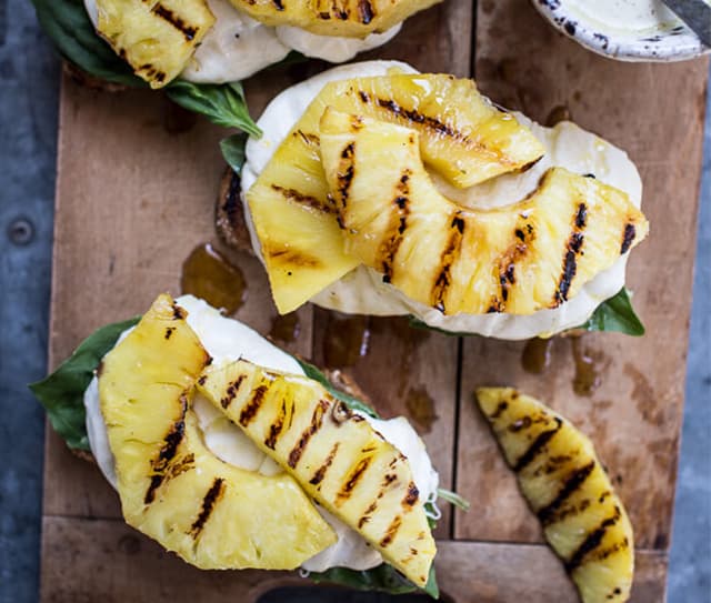 Grilled pineapple slices served on bread with mozzarella cheese and fresh basil, arranged on a wooden board.