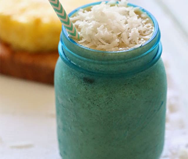 Blue mason jar filled with a green smoothie, topped with shredded coconut and a straw; toast with pineapple in the background.