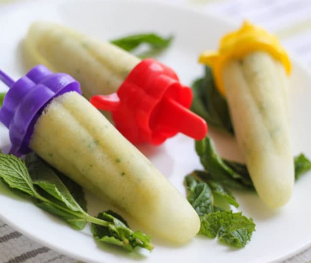 Three pale green popsicles with colorful handles rest on a white plate, garnished with fresh mint leaves.