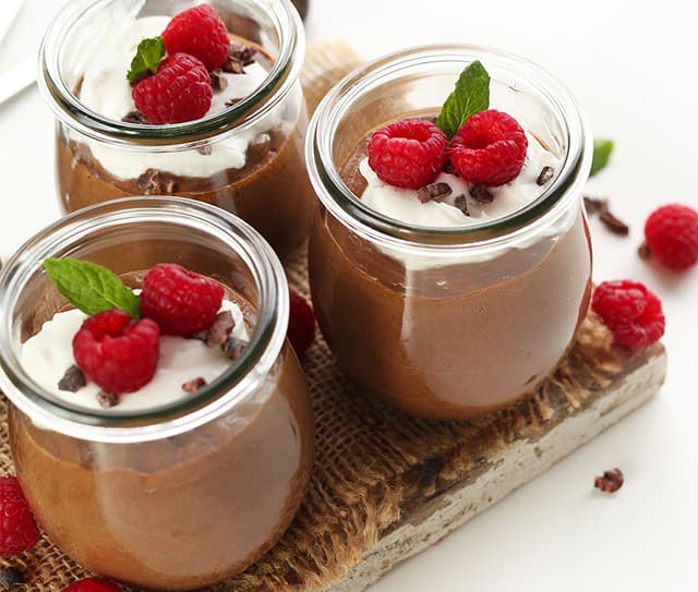 Three small glass jars filled with chocolate mousse, topped with whipped cream, raspberries, cacao nibs, and mint leaves, arranged on a rustic wooden board.