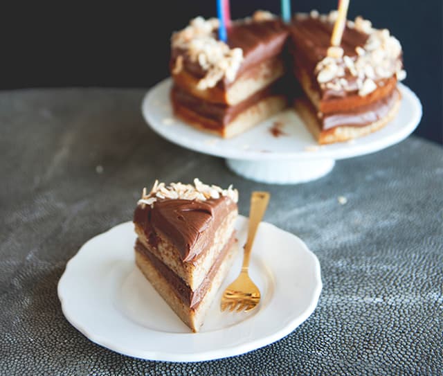 Slice of layered chocolate and vanilla cake with frosting on a white plate, gold fork beside it; whole cake with missing slice on a cake stand in the background.