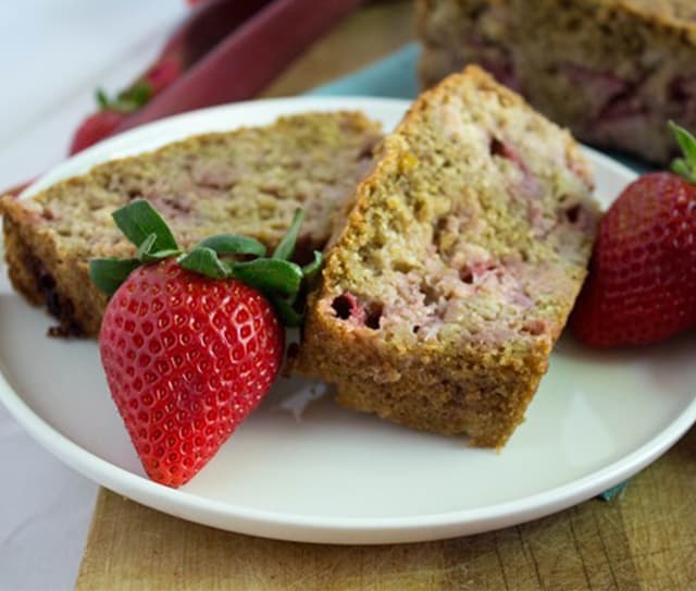 Two slices of strawberry bread on a white plate, garnished with two whole strawberries, with additional bread in the background.