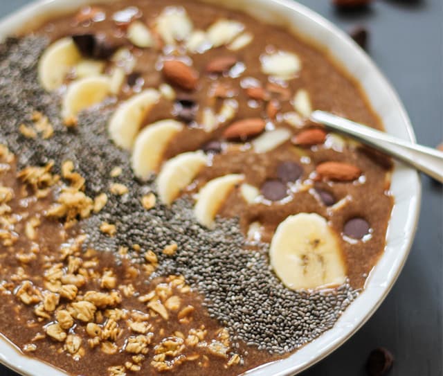 A bowl of chocolate smoothie topped with banana slices, chia seeds, granola, almonds, and chocolate chips, with a spoon on the side.