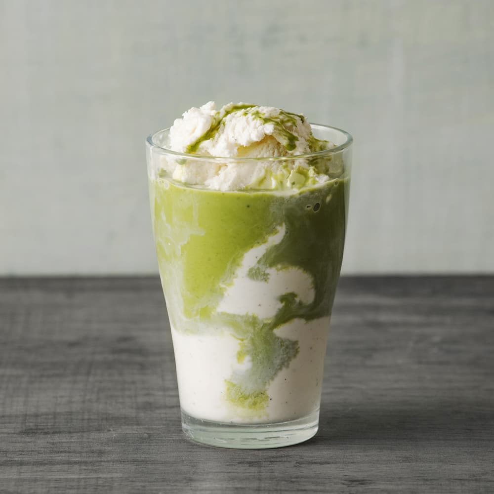 A glass filled with layers of green matcha and white ice cream, set on a gray surface with a light gray background.