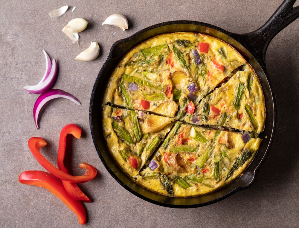 A vegetable frittata in a cast iron skillet is surrounded by sliced red pepper, red onion, and garlic cloves on a gray surface.