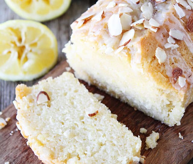 A sliced loaf of lemon almond bread topped with glaze and almond slices, placed on a wooden surface with lemon halves in the background.