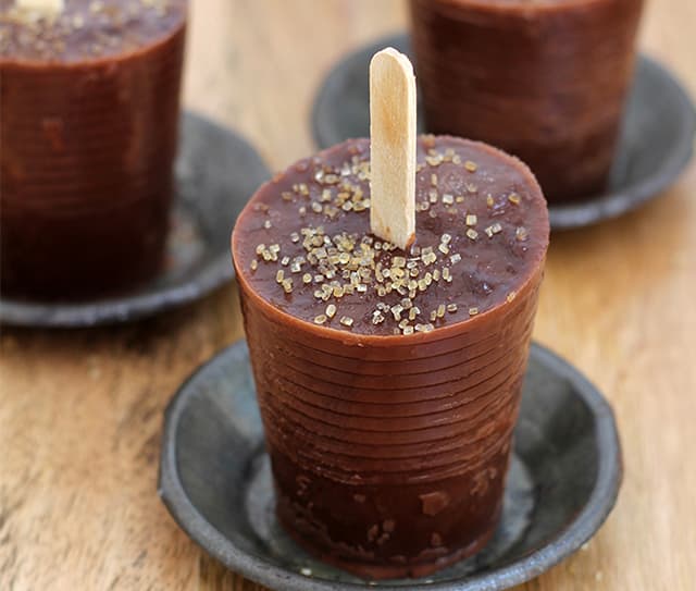 Three chocolate ice pops in cylindrical molds with wooden sticks, topped with coarse sugar, are placed on small round plates on a wooden surface.