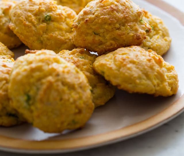 A plate of golden, round drop biscuits with a slightly rough texture, arranged in a casual pile.