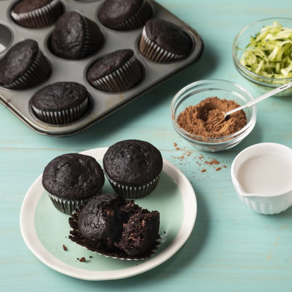 A plate with three chocolate muffins, one partially eaten, sits next to a muffin tray, bowls of cocoa powder and shredded zucchini, and a small bowl of milk on a blue surface.