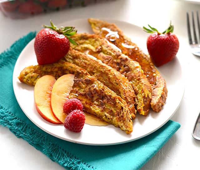 Plate with three slices of French toast, topped with syrup, served with two strawberries, peach slices, and raspberries on a white plate with a teal napkin.