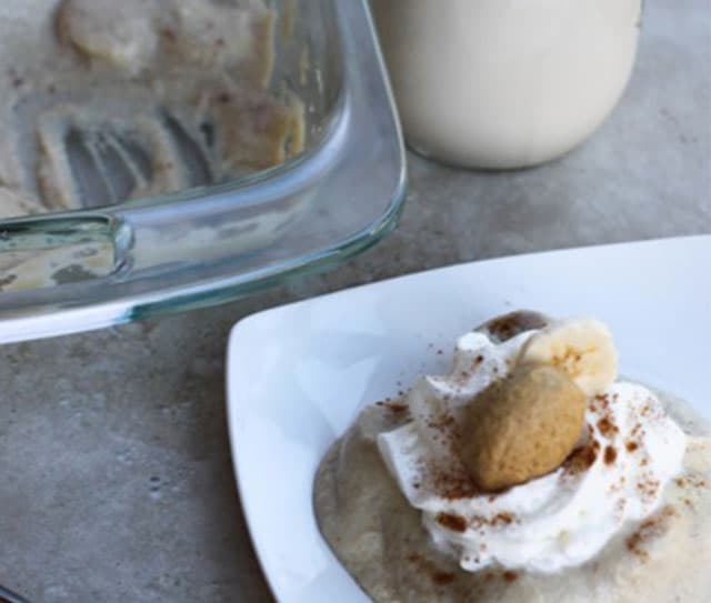 A dessert with whipped cream, banana slice, and a cookie on a plate; an empty glass baking dish and a jar are in the background.