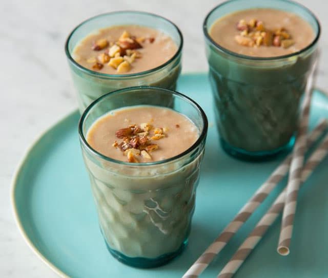 Three glasses of creamy smoothie topped with chopped nuts are on a blue tray, with two striped straws beside them.