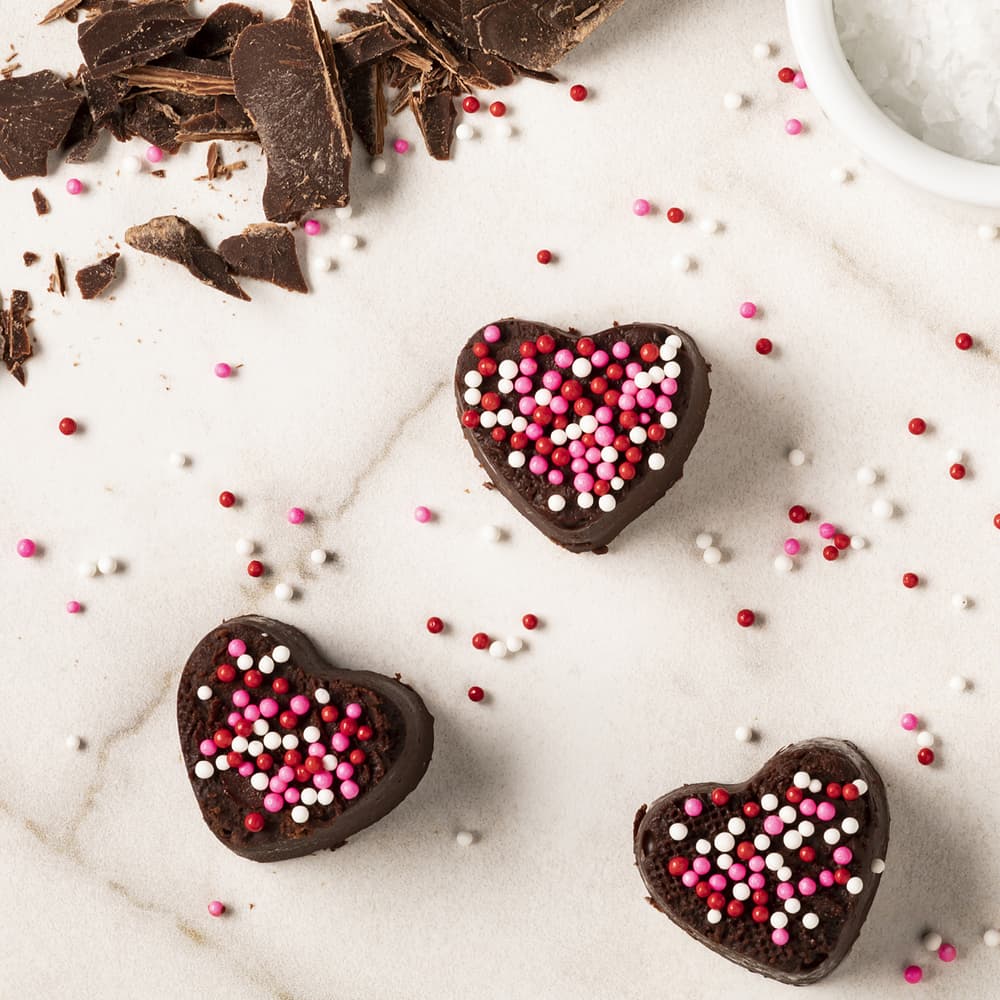 Three heart-shaped chocolate treats with pink, white, and red sprinkles on a marble surface, surrounded by chocolate pieces and scattered sprinkles.