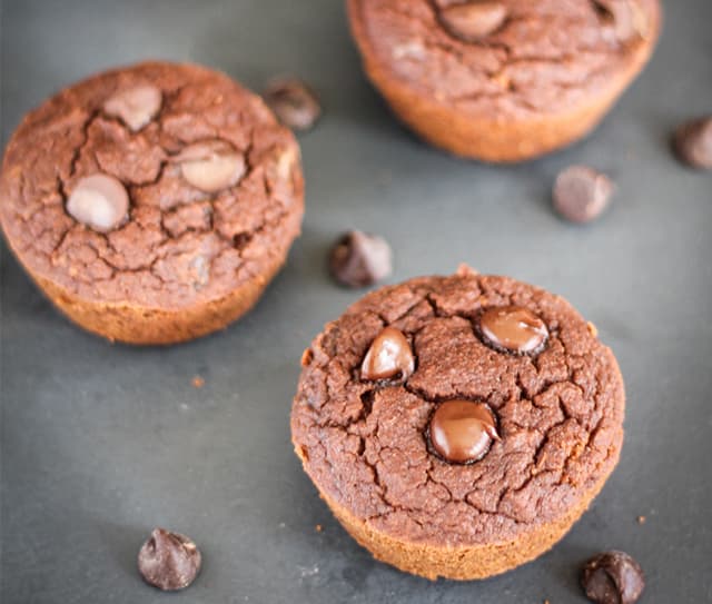 Three chocolate muffins with chocolate chips on top are placed on a dark surface, with a few loose chocolate chips nearby.