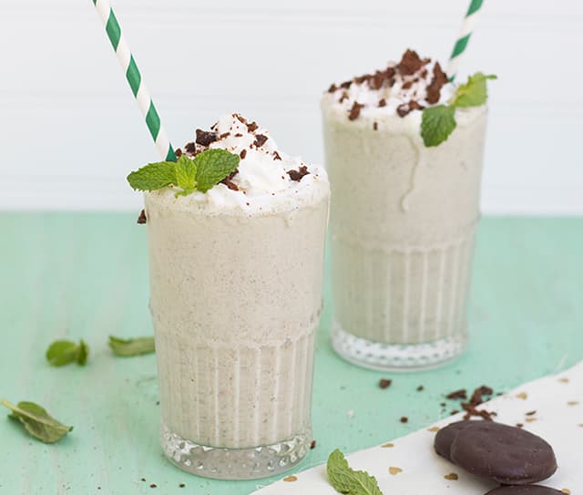 Two glasses of mint chocolate chip milkshake topped with whipped cream, crushed cookies, and a sprig of mint, each with a striped straw. Cookies and mint leaves are scattered nearby.