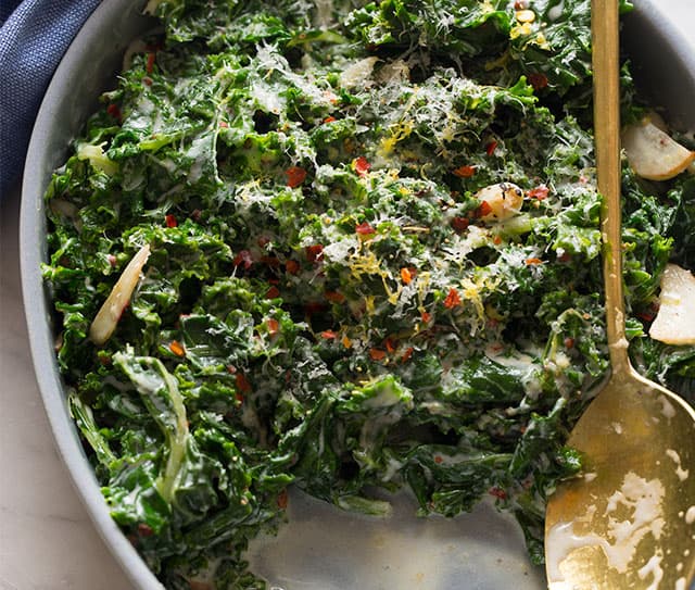 Cooked kale with creamy sauce, sliced garlic, grated cheese, and red pepper flakes in a round serving dish with a gold serving spoon.