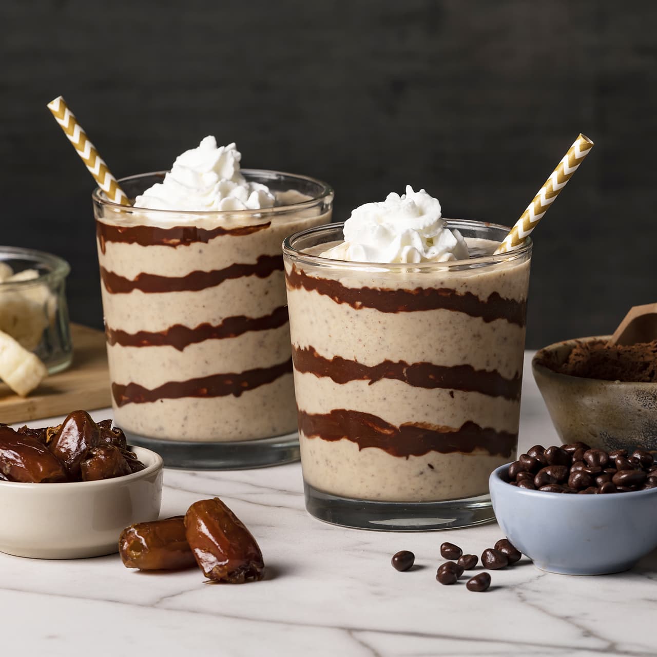 Two glasses of blended smoothie with chocolate stripes, topped with whipped cream and straws. Bowls of dates, bananas, and coffee beans are on the side.