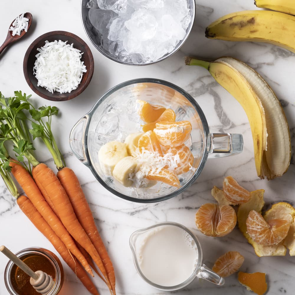 A blender with ice, banana slices, orange segments, and shredded coconut surrounded by carrots, bananas, tangerines, shredded coconut, a bowl of ice, honey, and a jug of milk on a marble surface.