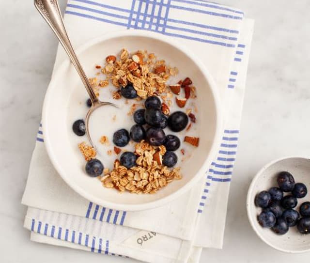 A bowl of granola, blueberries, and milk with a spoon, placed on a napkin next to a small bowl of blueberries.