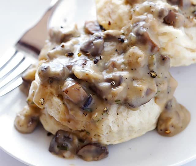 A biscuit topped with creamy mushroom gravy and black pepper sits on a white plate next to a fork.