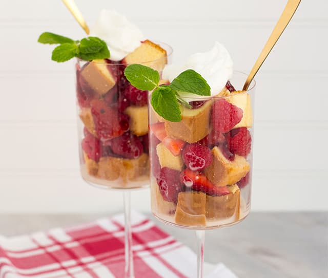 Two glasses filled with cubes of pound cake, fresh berries, whipped cream, and mint leaves, with gold spoons, set on a red and white cloth.