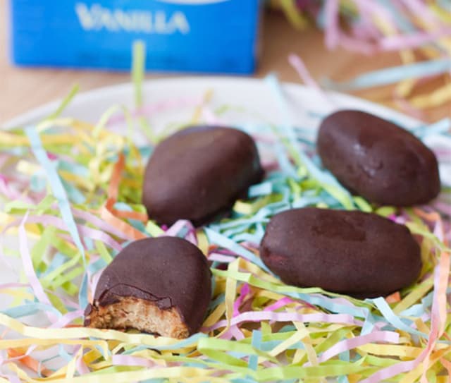 Four chocolate-covered treats sit on a bed of colorful shredded paper, with one treat showing a bite revealing a light-colored filling. A blue box is partially visible in the background.