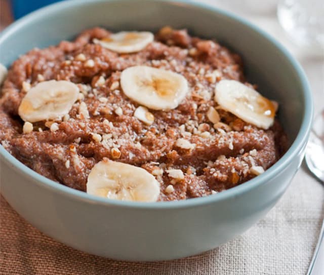 A bowl of chocolate oatmeal topped with banana slices and chopped nuts sits on a table next to a spoon.