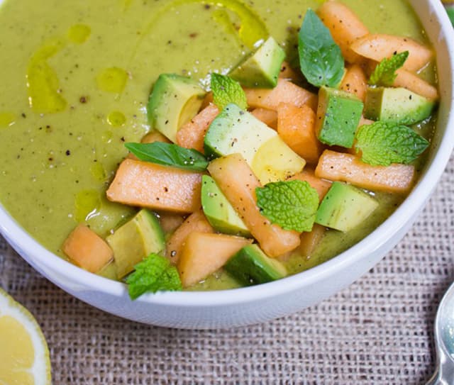 A bowl of green soup topped with diced avocado, cantaloupe, fresh mint leaves, black pepper, and a drizzle of olive oil.