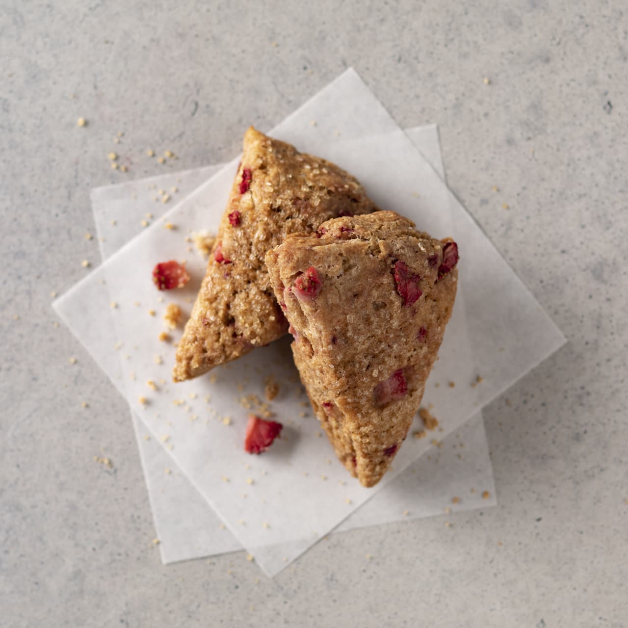 Two triangular scones with visible pieces of dried strawberries, placed on parchment paper on a light-colored surface, with crumbs scattered around.